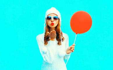Happy young woman blowing red lips sends air kiss holding air balloon in white knitted hat on colorful blue wall background