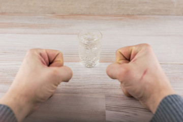 the struggle with addiction - alcoholism: a shot of vodka on the table in front of a man, clenched fists, in search of sufficient strength of will to resist temptation