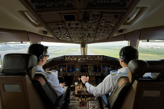 Two airliner pilots are flying the airplane towards the runway. Outside cockpit can see landing runway and airport. Inside cockpit can see pilots and all flight instrument and equipment.