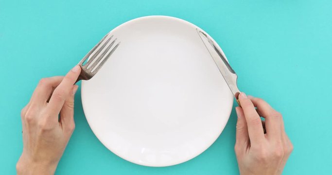 Female hands with cutlery and empty plate on turquoise background. Top view of nervous hungry female hands at dinner table holding fork and a knife above empty flat white plate