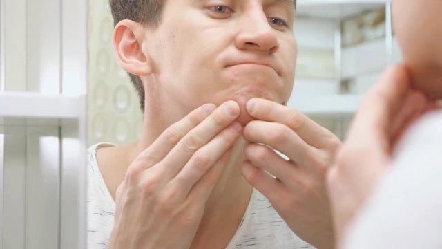 close up of young man standing close to a mirror in the bathroom and squeezing a pimple on his face.