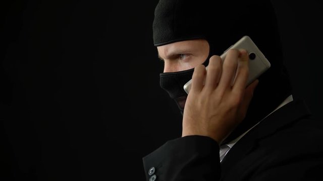 Killer in balaclava talking on phone, holding gun and planning crime, corruption