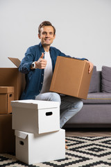 Image of happy brunet sitting on floor with cardboard boxes on background of gray sofa