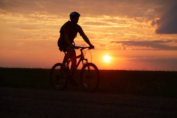 Fototapeta na wymiar Silhouette of a cyclist on the background of the sunset. Biker rides on the road in the evening. Red-yellow background outdoors