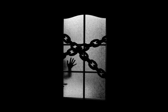 Dark silhouette of hand behind glass door with chain. Locked alone in room behind door on chain on Halloween. Night kidnapping. Evil in home on monochrome. Сhild inside haunted house in grayscale.