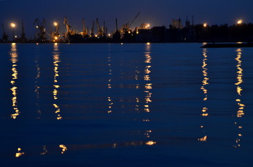 Sea port in the evening with the reflection of light from the lanterns on the water. Blue sea water with lines of light. Cranes, berth.