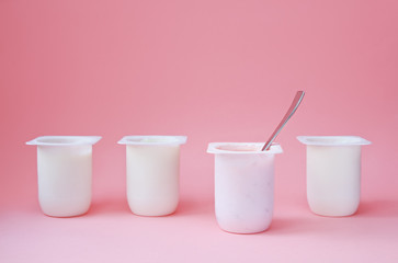 Four yogurts in white plastic cups on pink background with copy space. Strawberry pink yoghurt with spoon in it. Minimal style. Concept of better choice.