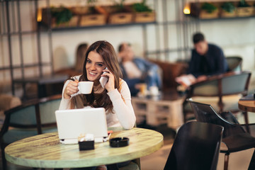 Young woman sitting in front of open laptop computer in cafe bar using phone