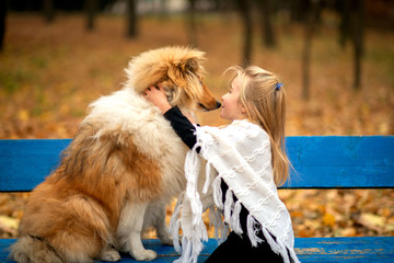 A girl in a white poncho is sitting on a bench in the autumn park and kiss a red collie dog