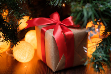 A gift in craft paper with a red ribbon on the background of a New Year's decorated Christmas tree with lights.