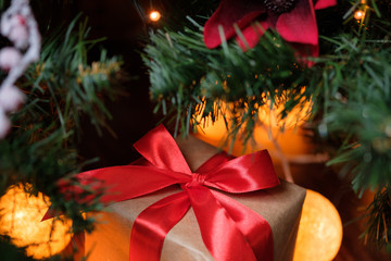 A gift in craft paper with a red ribbon on the background of a New Year's decorated Christmas tree with lights.
