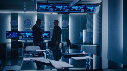 Federal Special Agent Talks To Military Man in the Monitoring Room. In the Background Busy System Control Center with Monitors Showing Data Flow.