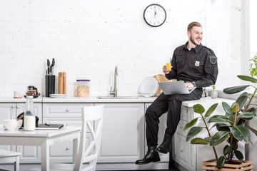 smiling man in police uniform sitting on kitchen table, drinking orange juice and using laptop