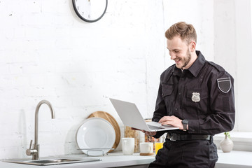 handsome man in police uniform smiling and using laptop at kitchen