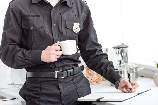 Police Officer Drinking Coffee And Writing In Notebook At Kitchen Table