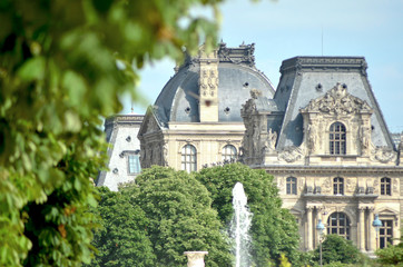 Fototapeta na wymiar The classical buildings of the Louvre are framed by the trees of the Tuileries Gardens. A fountain is in the foreground. The sky is blue.