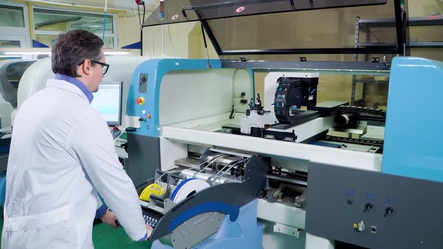 The engineer in a white robe and glasses works for the Surface Mount Technology machine. PCB Processing on CNC machine, Production of electronic components at high-tech factory