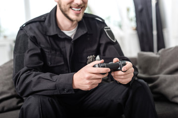 cropped view of smiling policeman with gamepad sitting on couch