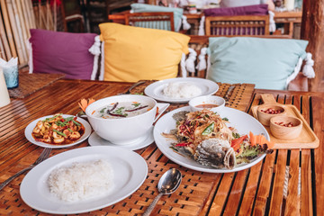 Thai dinner food with rice, Stir fried calamari and shrimp, Chicken soup with coconut milk, chicken grilled BBQ sauce, fried barramundi with herb on the white plate over wooden table in restaurant