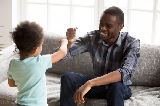 Happy African American smiling father playing with little toddler son in Rock Paper Scissors at home in sitting room, family spending time together, toddler playing with man sitting on couch