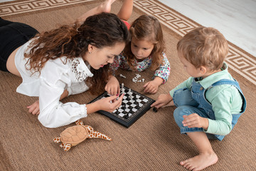 A young mother or nanny or older sister lies on the floor on the carpet at home with small young children and teaches them to play checkers or chess.