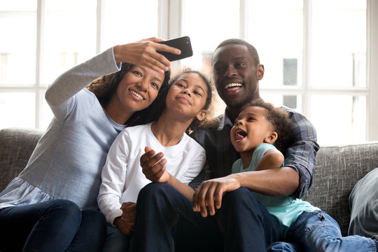Happy African American large family taking selfie on phone together, attractive mother holding smartphone phone, taking family photo, smiling father, little preschooler daughter and toddler son posing