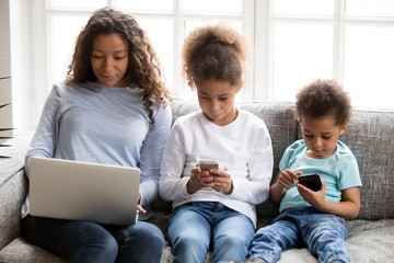 African American family using devices, ignoring each other, pretty mother using laptop, looking at screen, preschooler daughter and toddler son using phones, playing mobile apps, addicted with devices