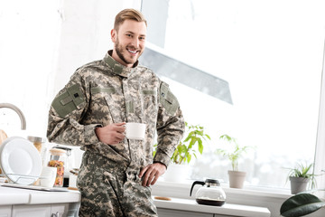 smiling army soldier holding cup of coffee in kitchen