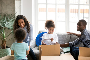 Fototapeta na wymiar Happy large African American family unpacking cardboard boxes with belongings, just moving, sitting together on couch, little daughter and toddler son helping parents with packaged things, new home