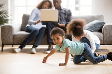 African American married couple using laptop together, sitting on couch at new home while children playing, toddler son playing with toy car on floor, little daughter drawing colorful pencil