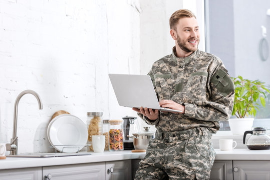 smiling army soldier using laptop at kitchen and looking away