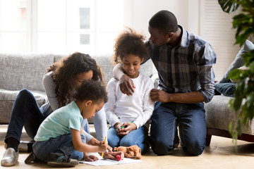 Large African American family playing together on warm floor in living room, mother help toddler son with drawing, loving father embracing little preschooler daughter, family spending time together