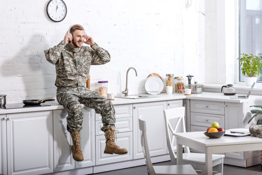 smiling soldier in headphones listening music and sitting on kitchen countertop at home