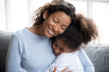 Loving African American mother embracing with preschooler little adorable daughter, sitting together on couch at home, warm relationships parent and child, closeness, love and support concept