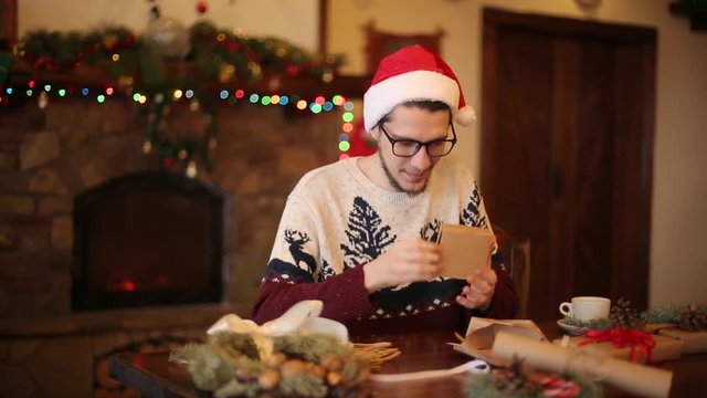 Happy man wearing santa hats opening Christmas gift box near fireplace with flashing garland. Smiling guy recieved a parcel with presents and enjoying unpacking package. Winter holidays concept.