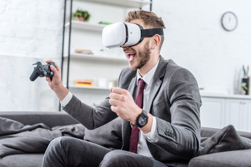 excited businessman wearing virtual reality headset and playing video game on couch at home