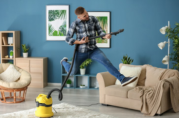 Young man having fun with vacuum cleaner while removing dirt in flat - obrazy, fototapety, plakaty