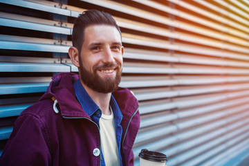 Handsome young man in purple winter jacket drinking coffee. The guy drinking coffee on the street. Sun flare