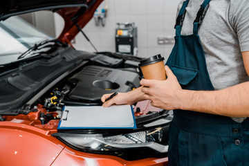 partial view of male auto mechanic with notepad and coffee to go near car at auto repair shop