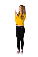 A full-length shot of a young girl with yellow sweater pointing with the index finger and looking up on isolated white background