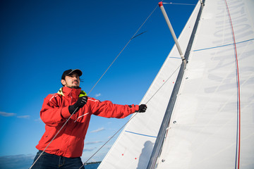 Young man skipper in red windbreaker and black cap sets the sails on a sailing yacht