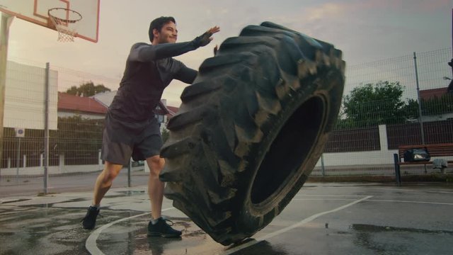 Strong Muscular Fit Young Man in Sport Outfit and Gloves is Doing Exercises in a Fenced Outdoor Basketball Court. He's Flipping a Big Heavy Tire in an Afternoon Environment After Rain.
