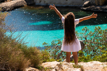 Open-armed girl with long hair watching the crystal clear water of Mediterranean sea in Mallorca.