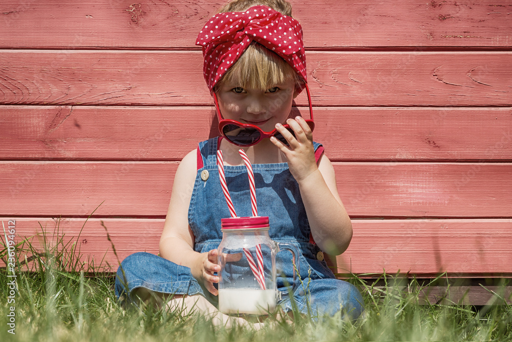 Wall mural Little girl in dungarees drinks milk from a jar. Red background, copy space.