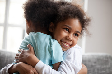 Smiling African American little girl embrace adorable brother, smiling preschooler sister hug cute toddler boy with love, sitting on couch together, good relations between siblings, close up