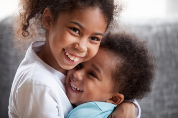 Obraz premium Happy African American siblings embracing, sitting together on couch at home in living room, little preschooler girl hug toddler adorable boy, good relations between sister and brother, close up