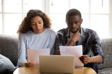 Serious focused African American couple reading paper documents, sitting together on couch at home, man and woman checking bills, bank account balance, terms of contract, mortgage, loan agreement