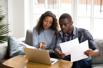 Serious African American couple discussing paper documents, sitting together on couch at home, man and woman checking bills, bank account balance, terms of contract, mortgage, loan agreement - Powered by Adobe