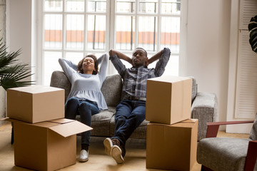 Calm African American couple in love relaxing leaning back on couch, sitting together, tired resting man and woman just arrived, unpacked cardboard boxes with belongings, moving concept, new house