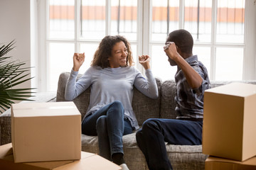 Happy African American couple in love celebrating moving in new house, sitting together on couch with raised hands, cardboard boxes with belongings, start living together, homeowners in new apartment